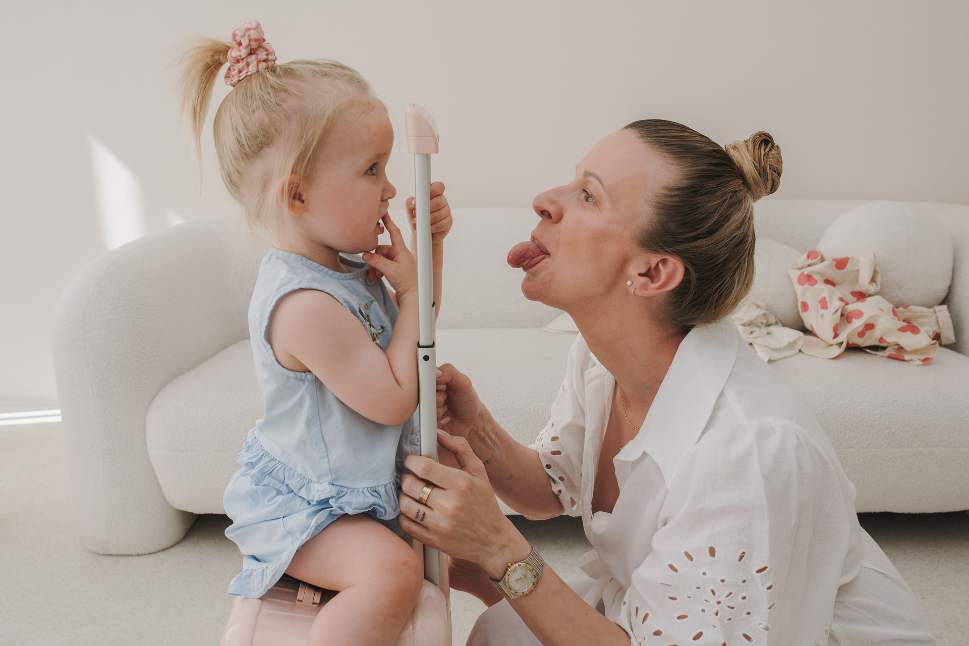 Woman playfully sticking out her tongue at a small child sitting on a suitcase handle indoors.