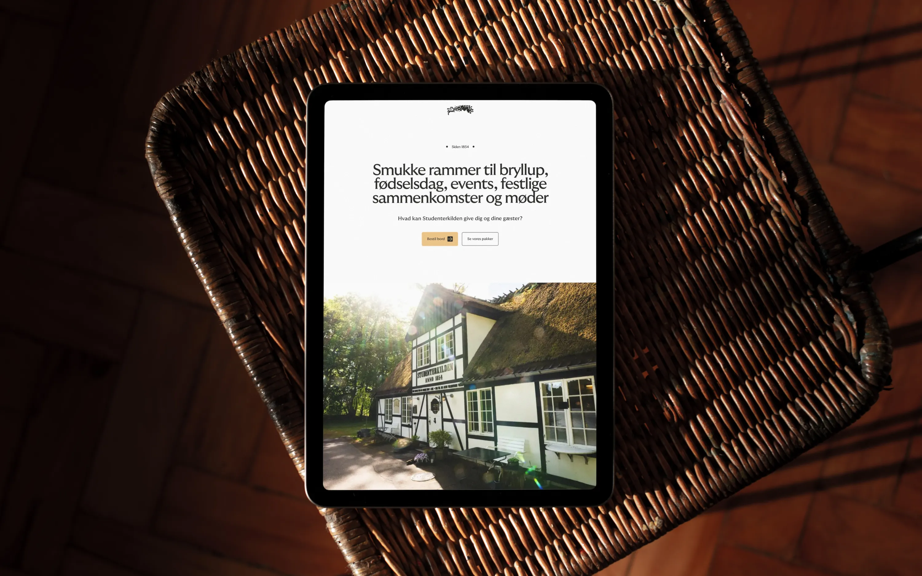 Tablet on a woven wicker chair displaying a webpage with Danish text about beautiful settings for weddings, birthdays, events, and meetings, with a photo of a traditional white and black timber house with a thatched roof and sunlight filtering through trees.