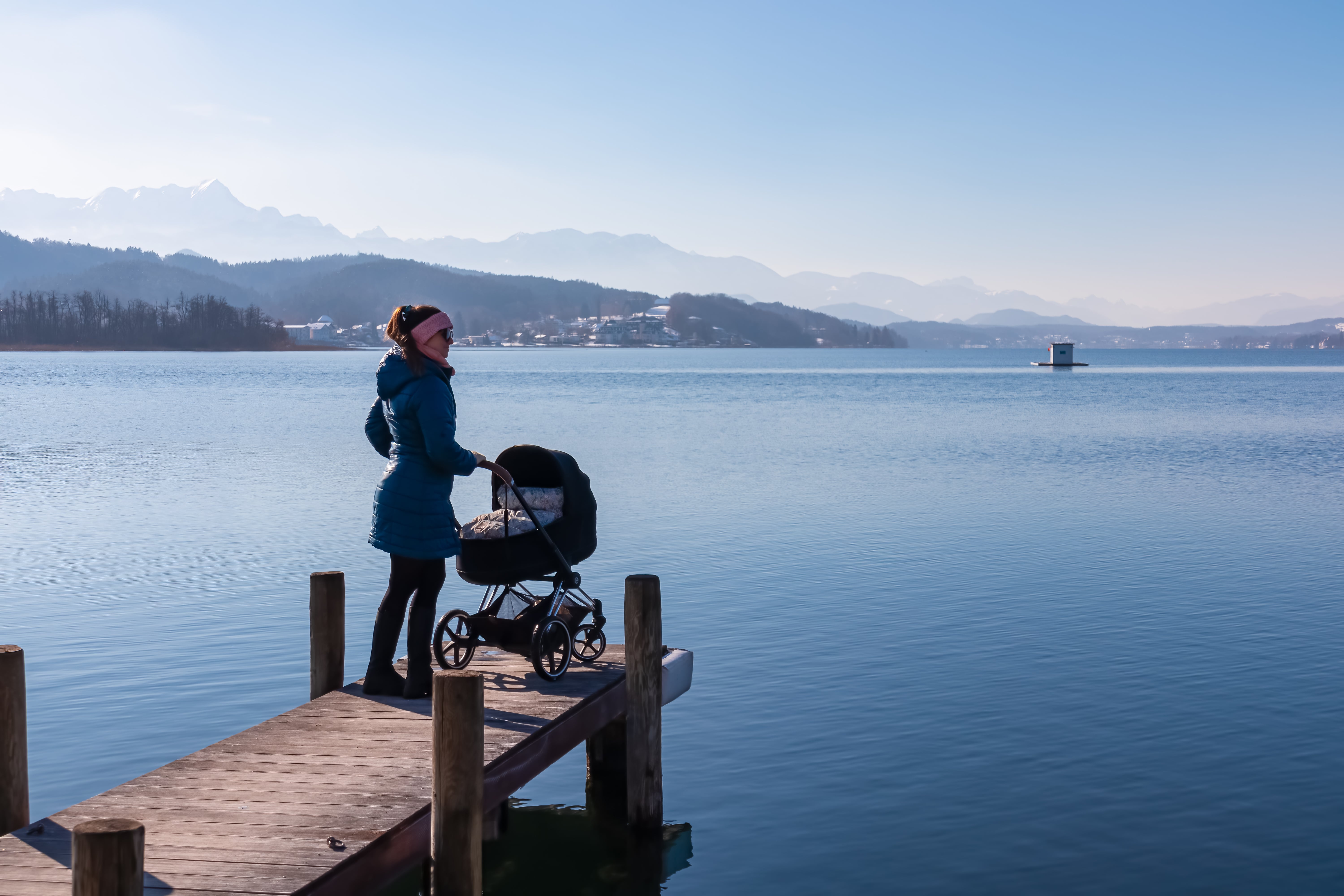 Woman with baby stroller standing on pier at idyllic Lake Wörth in Pörtschach, Carinthia, Austria. Scenic landscape surrounded by Karawanks, Austrian Alps. Fresh and clean air. Calm sunny winter day