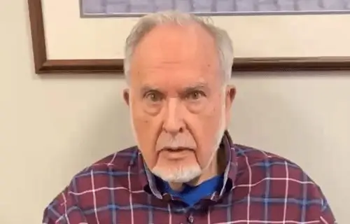 An older man with a white beard is sitting, wearing a colorful plaid shirt.