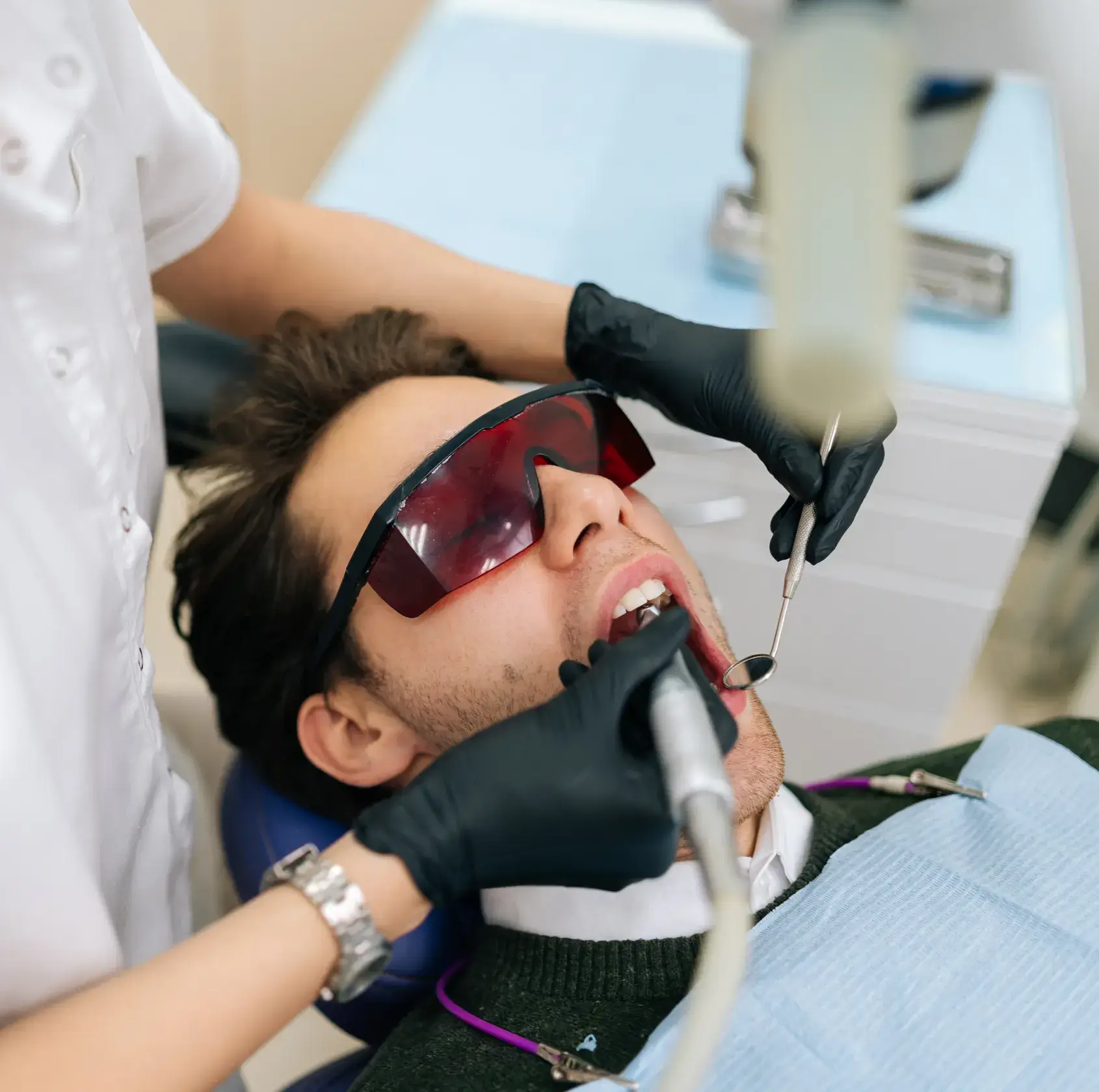 Person wearing protective glasses receiving dental treatment from a dentist using tools.