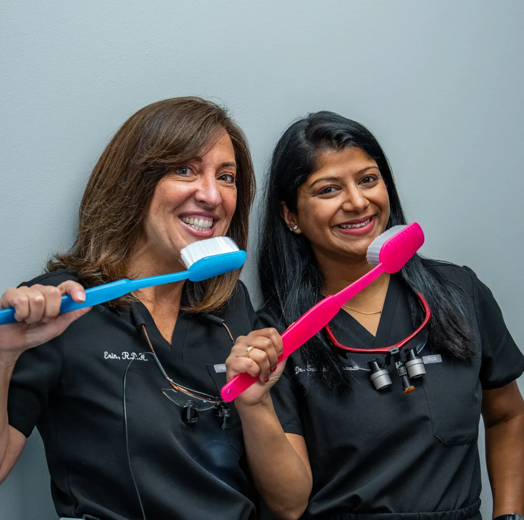Two women in dental scrubs smiling and holding oversized toothbrushes in front of a plain background.
