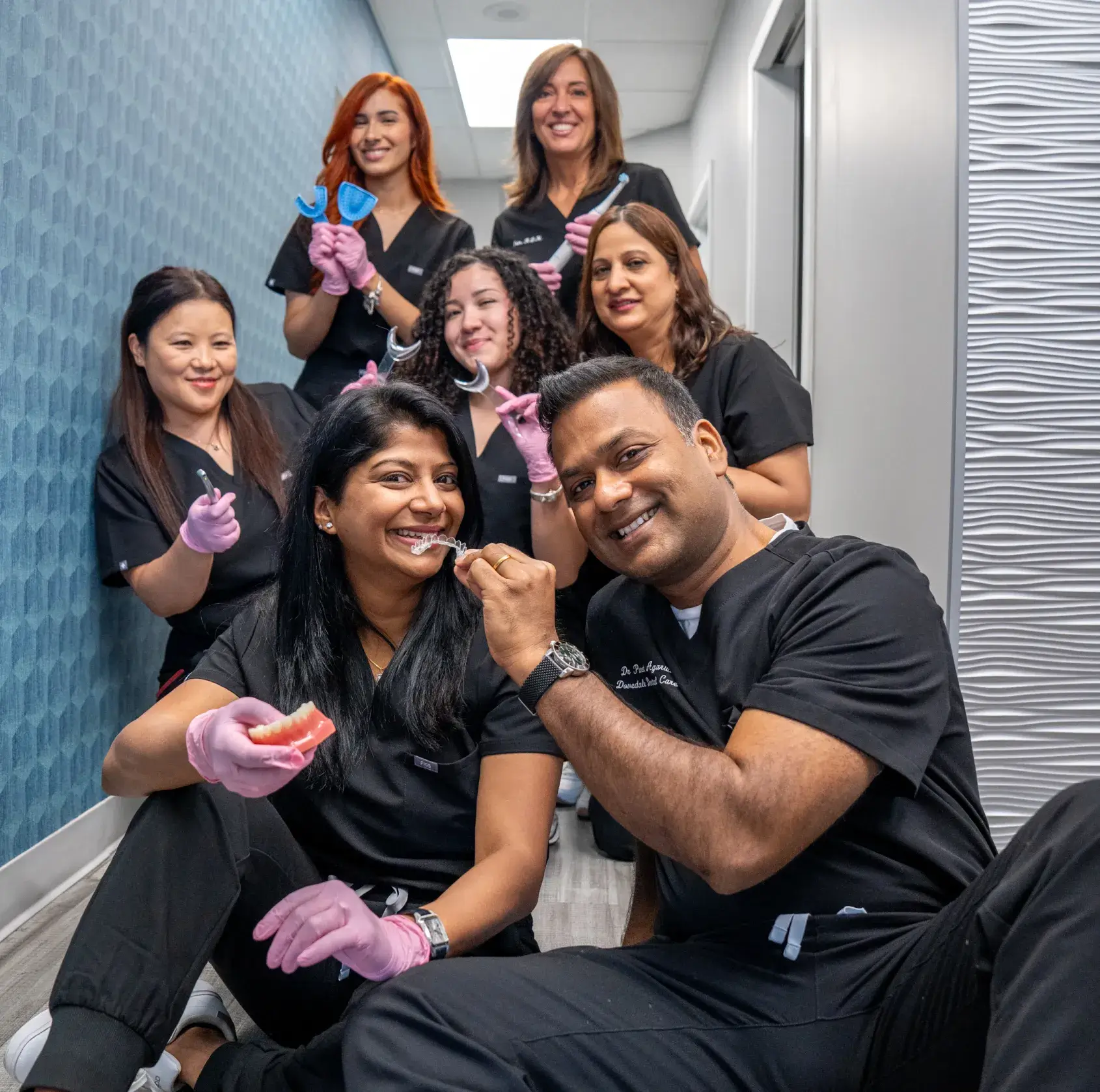 A group of dental staff pose playfully in a clinic hallway, holding dental tools and models.