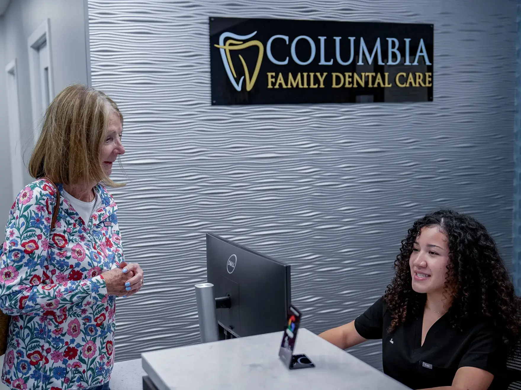 A person with curly hair smiles with arms crossed, standing in front of a textured wall.