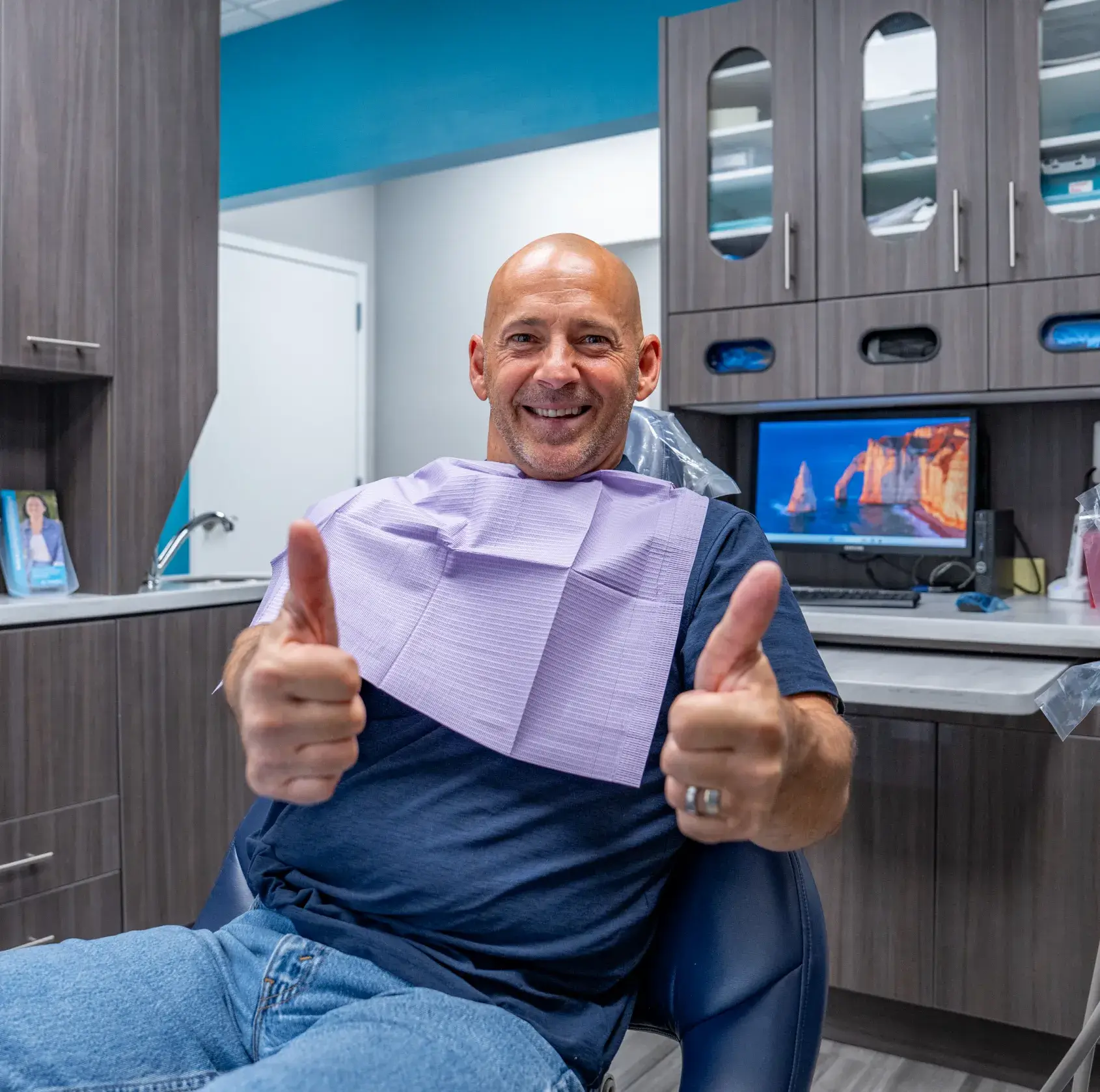 A man in a dental office gives two thumbs up while sitting in a chair.