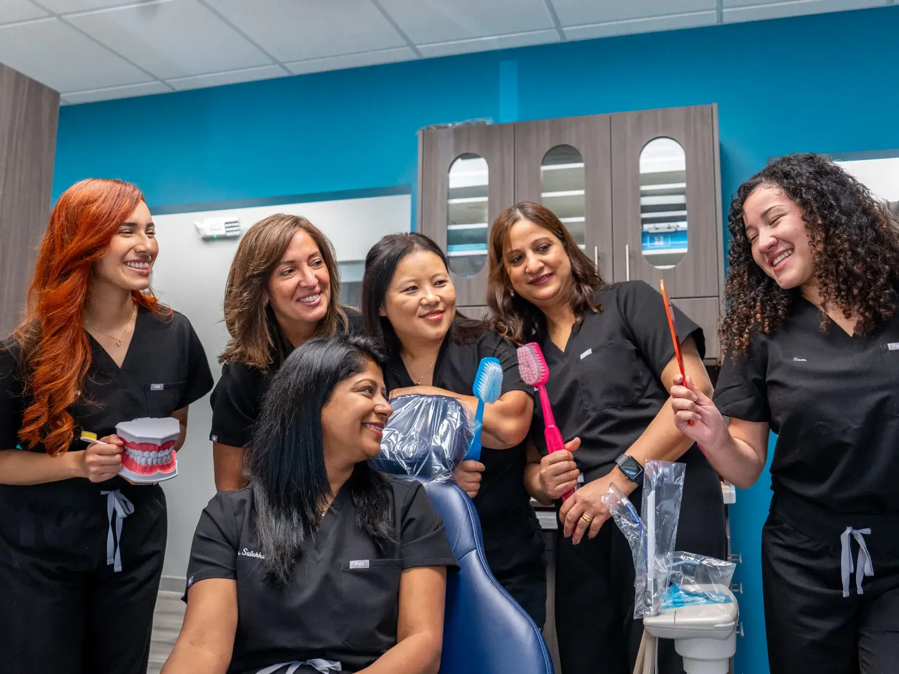 A group of six dental professionals in scrubs hold dental tools and a model, smiling together.