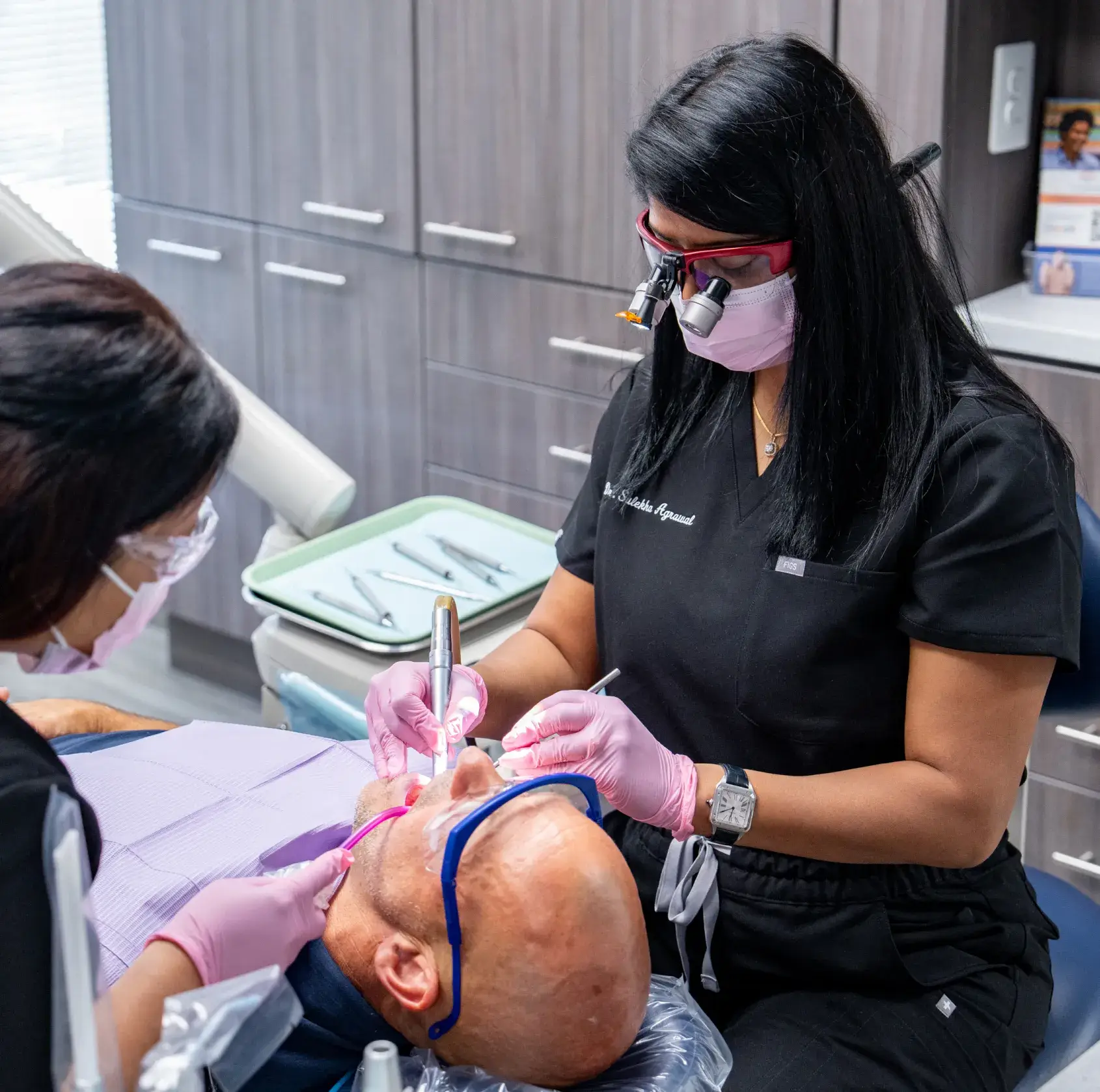 Two dentists in black scrubs discuss dental X-rays with a seated patient in a clinic.