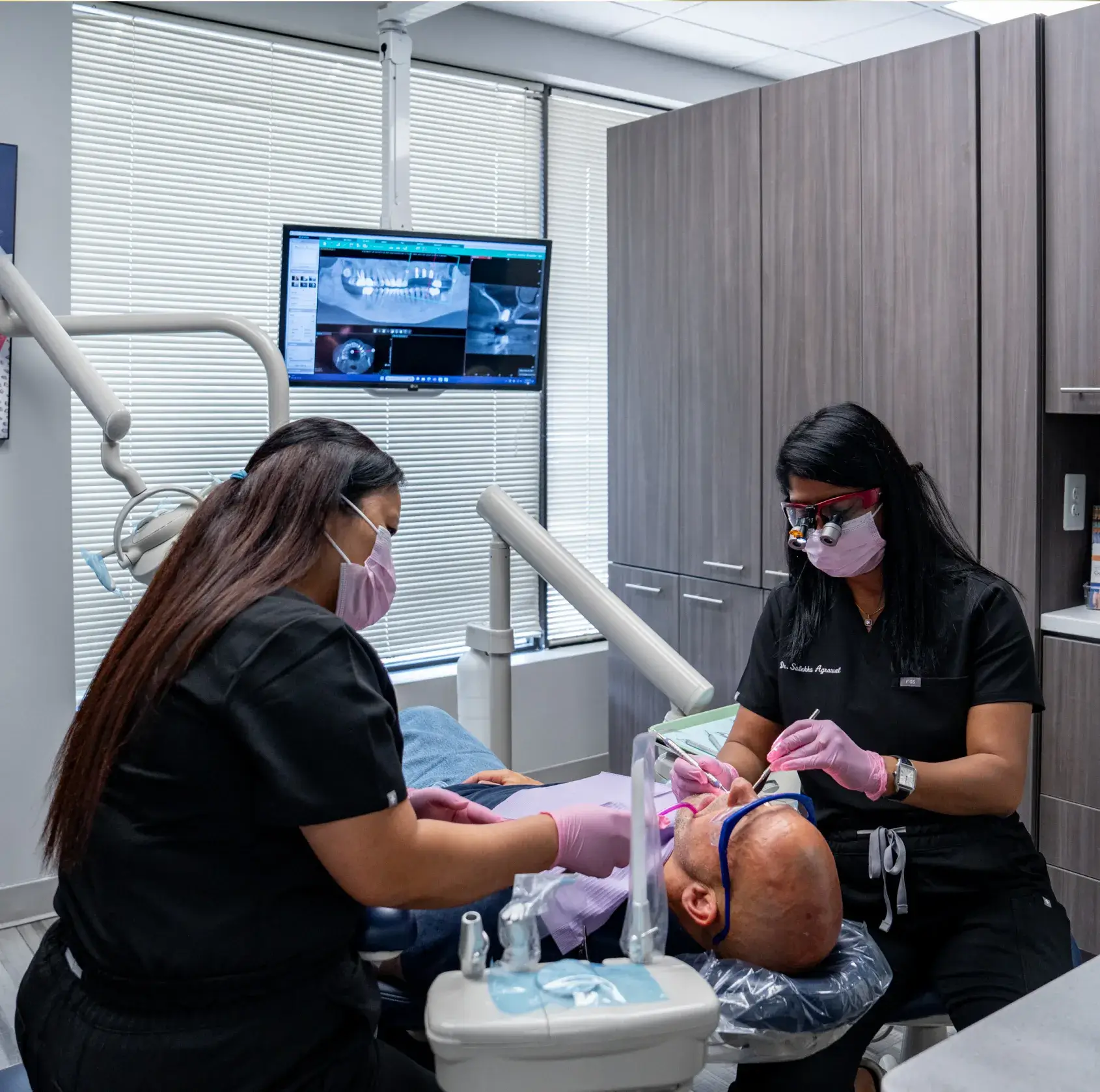 Two dental professionals work on a patient lying in a dental chair, with equipment nearby.