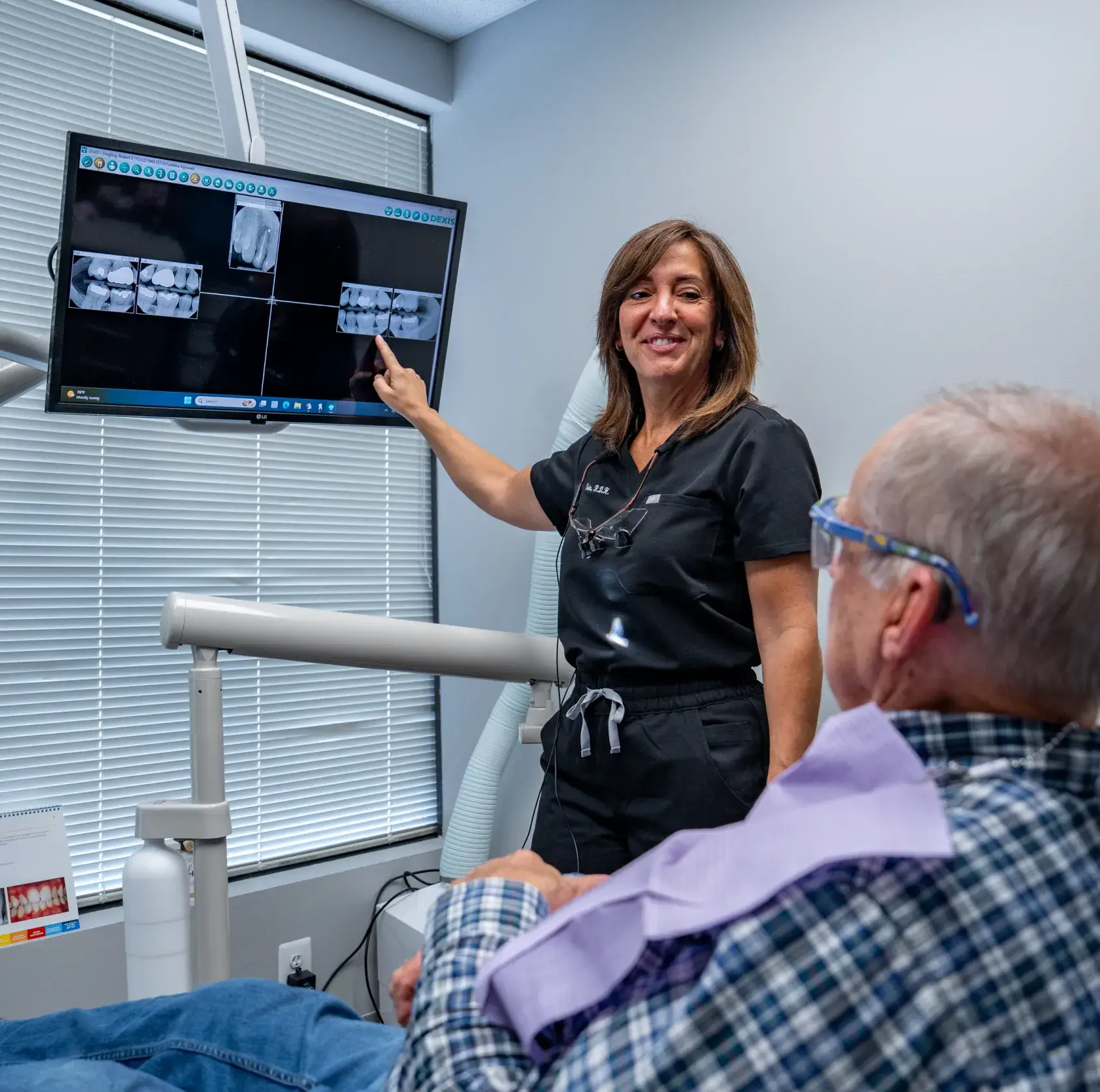 Two dental professionals discuss an X-ray with a seated patient in an exam room.