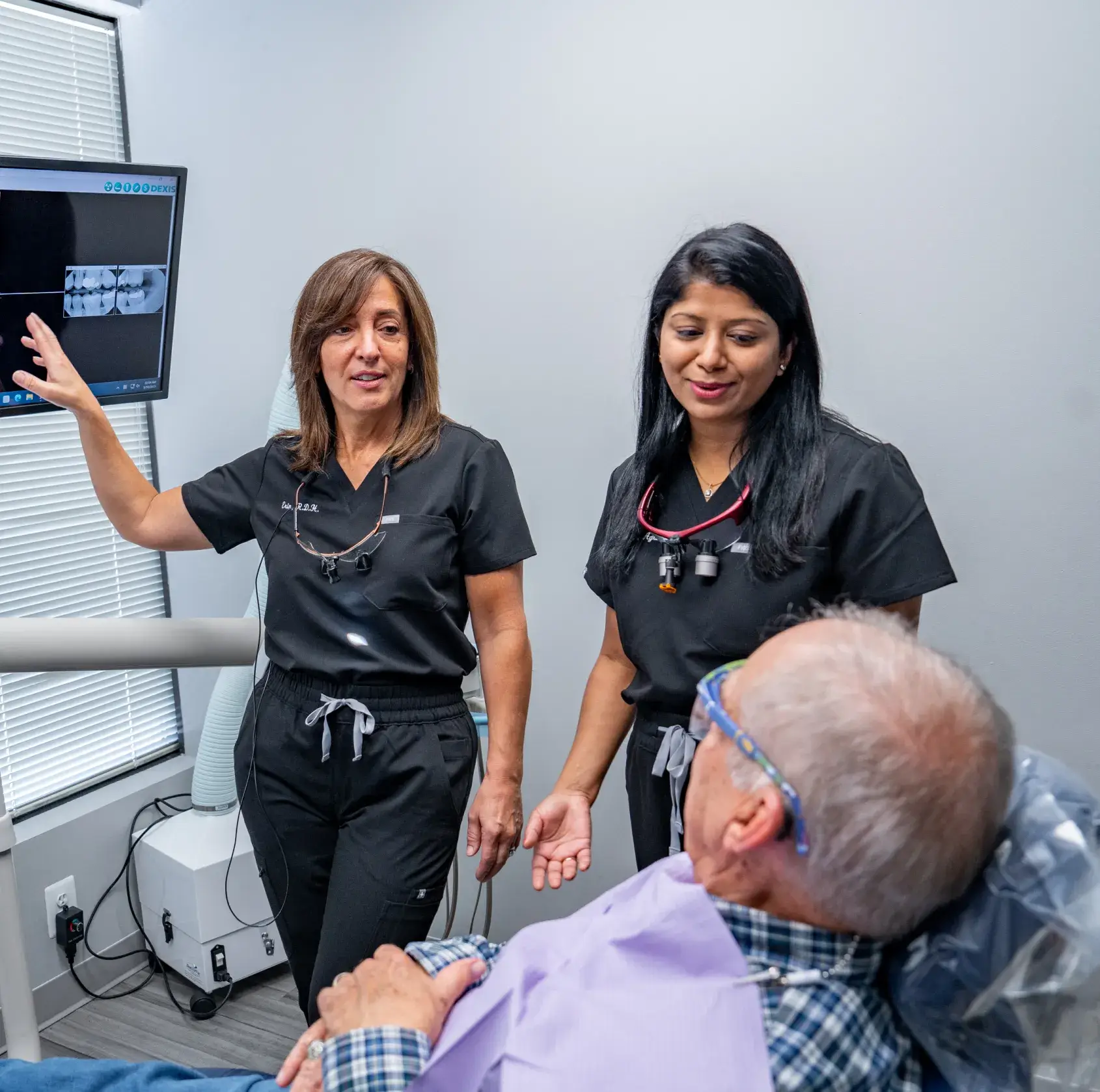 Two dental professionals discuss an X-ray with a seated patient in an examination room.