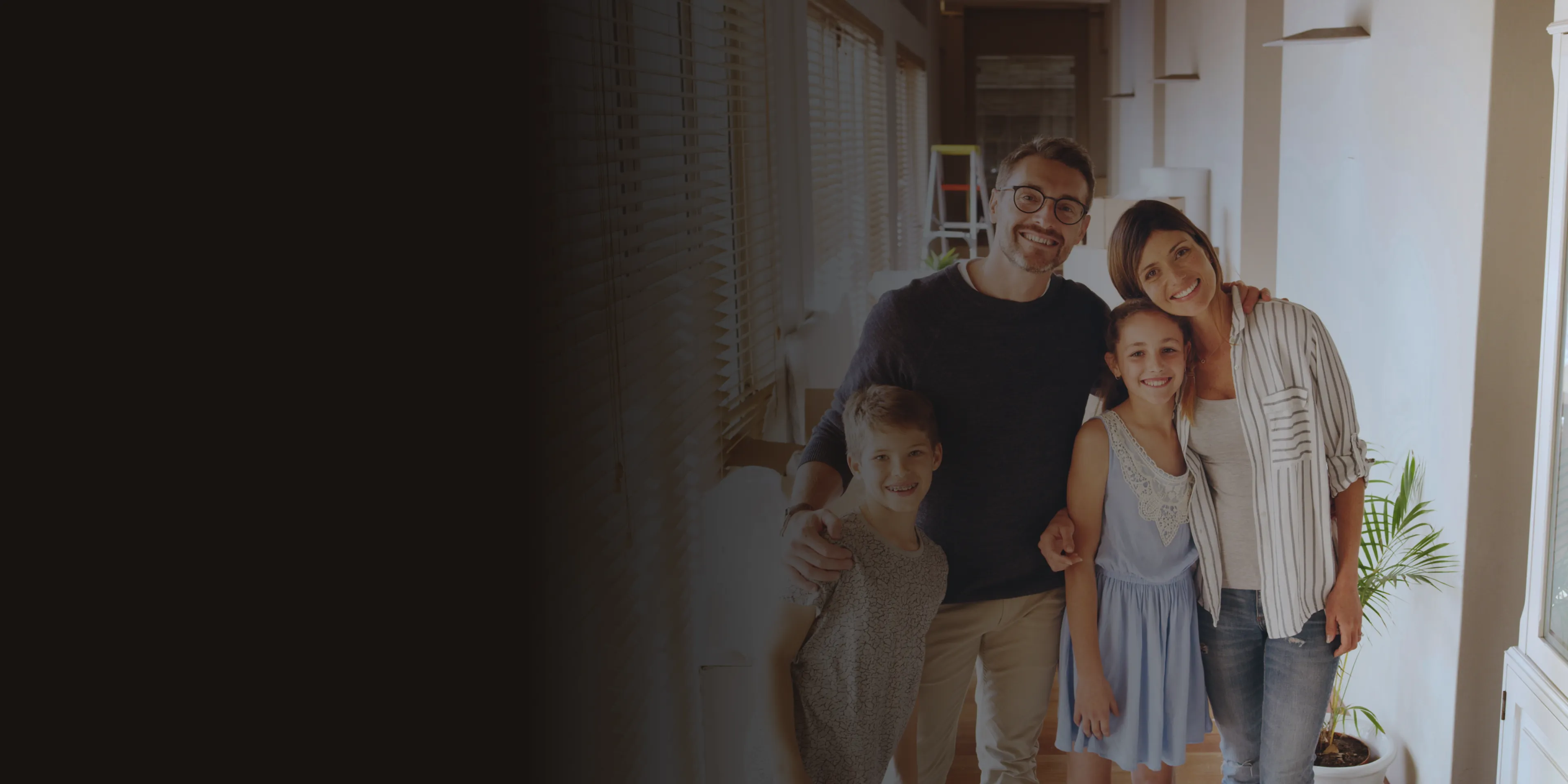 A family of four stands together, smiling, inside a well-lit room.