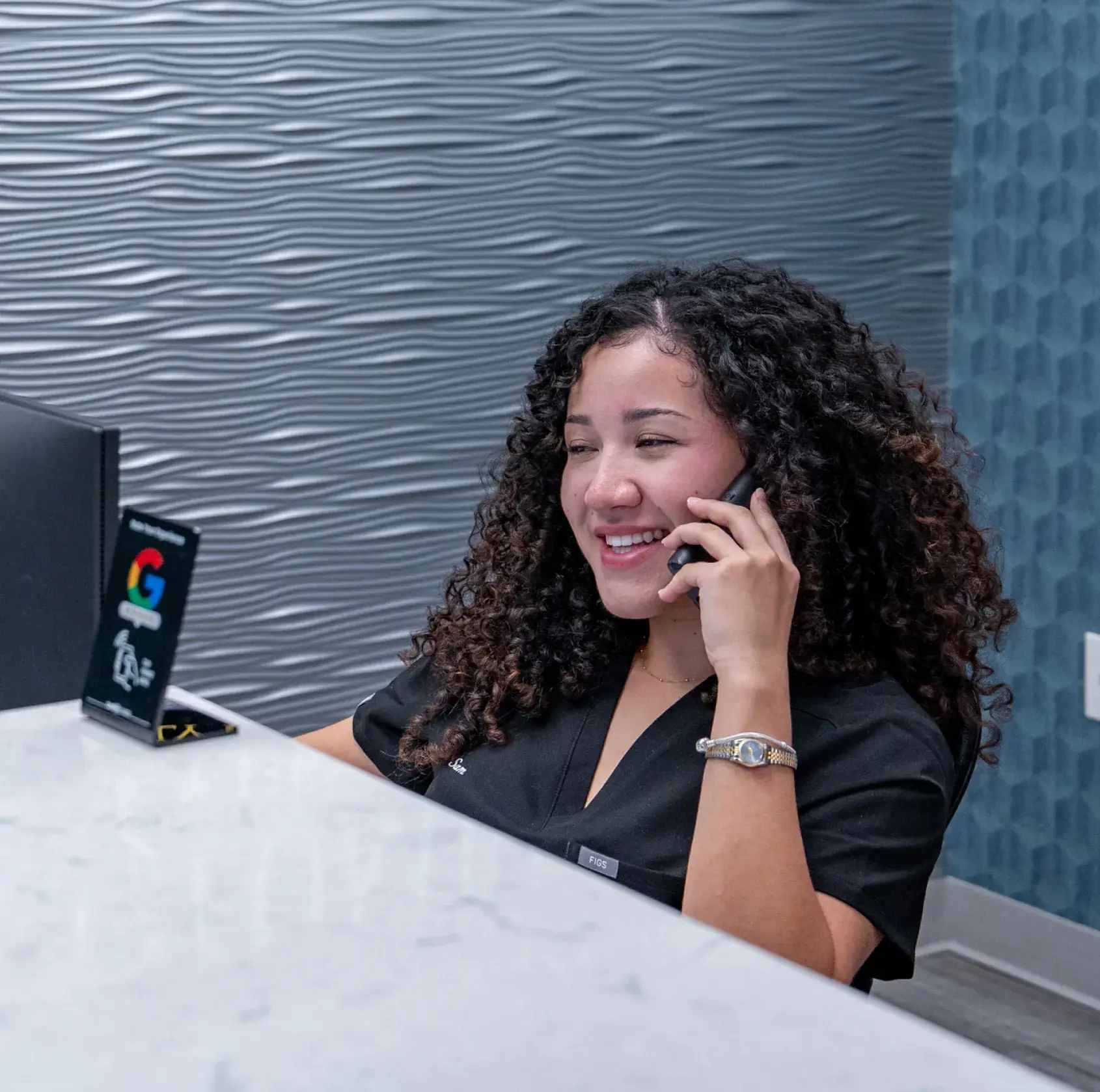 A woman with curly hair sits at a desk, smiling while talking on the phone.