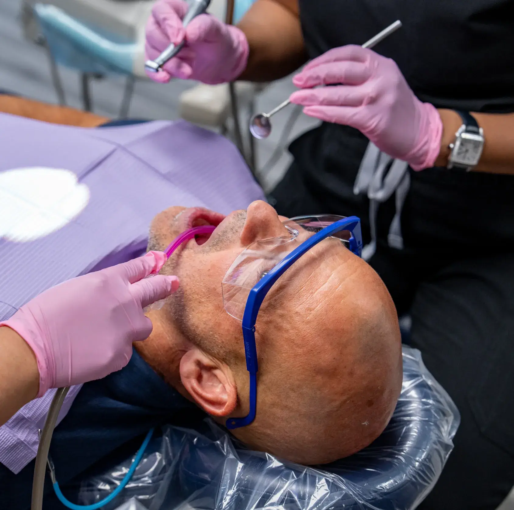 A dentist examines a patient lying back in the chair, using dental tools and wearing protective glasses.