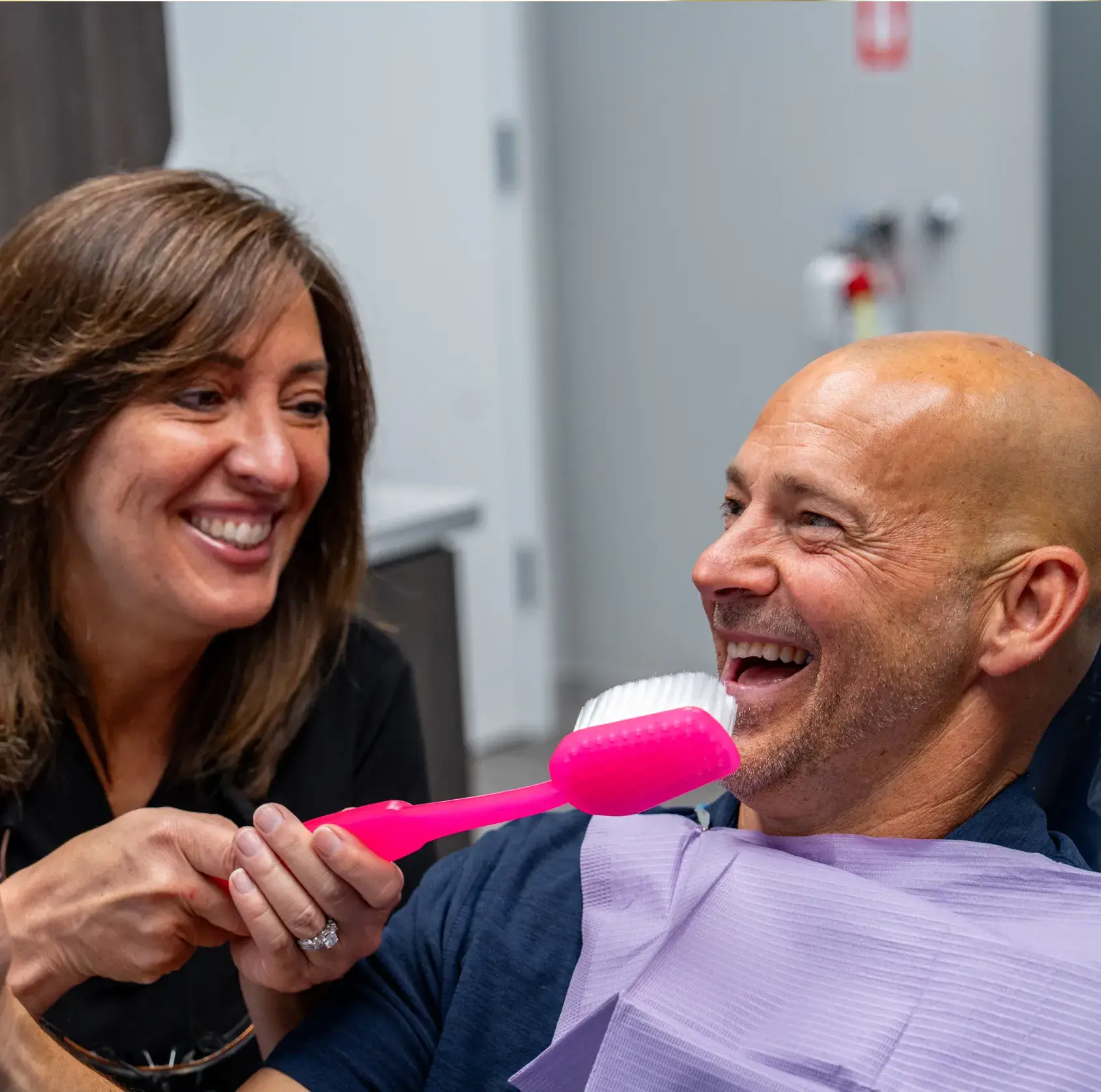 A woman uses a large pink toothbrush to humorously brush a man's teeth, both are smiling.