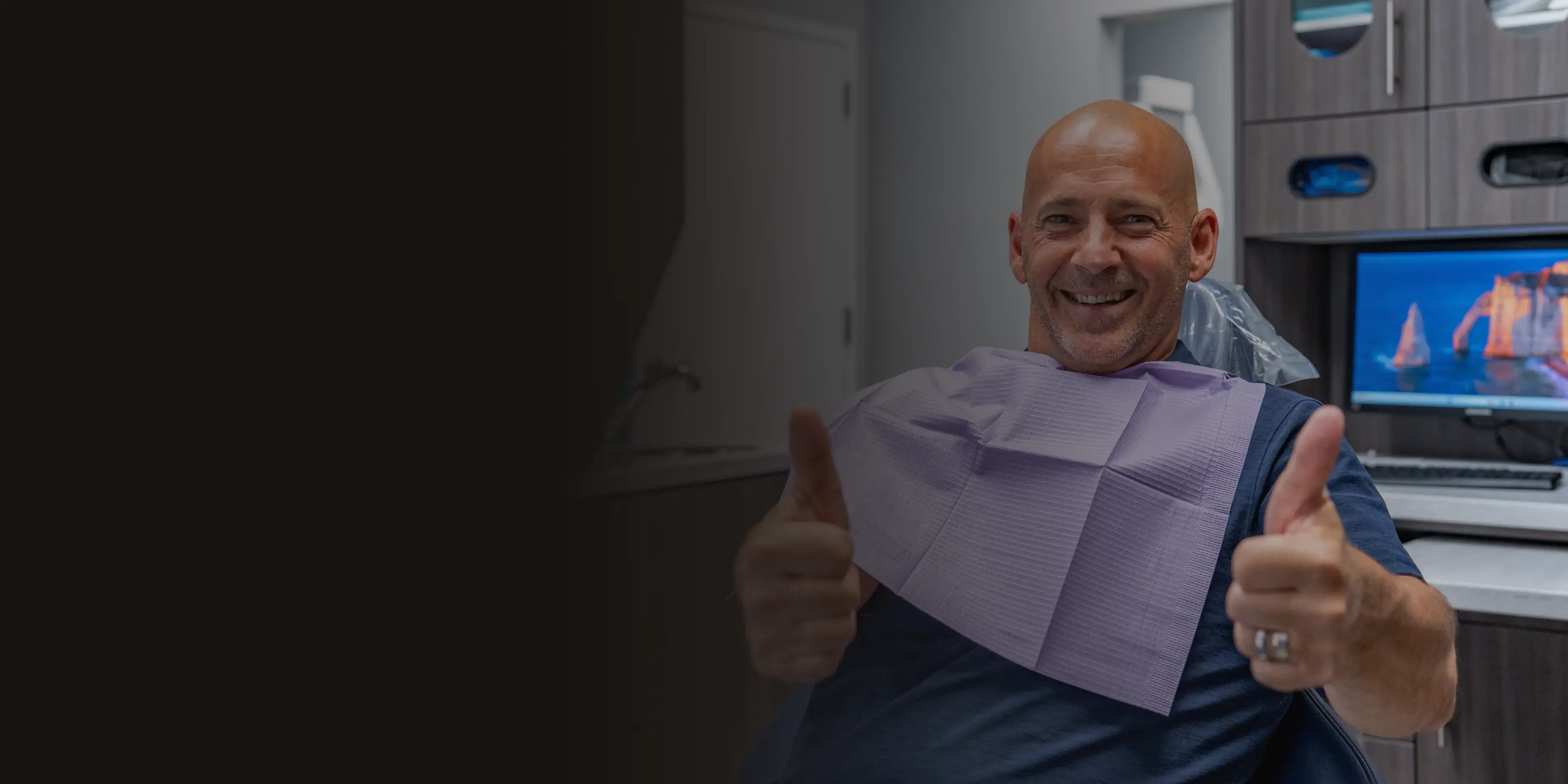 A man sitting in a dental chair gives a thumbs-up and smiles.