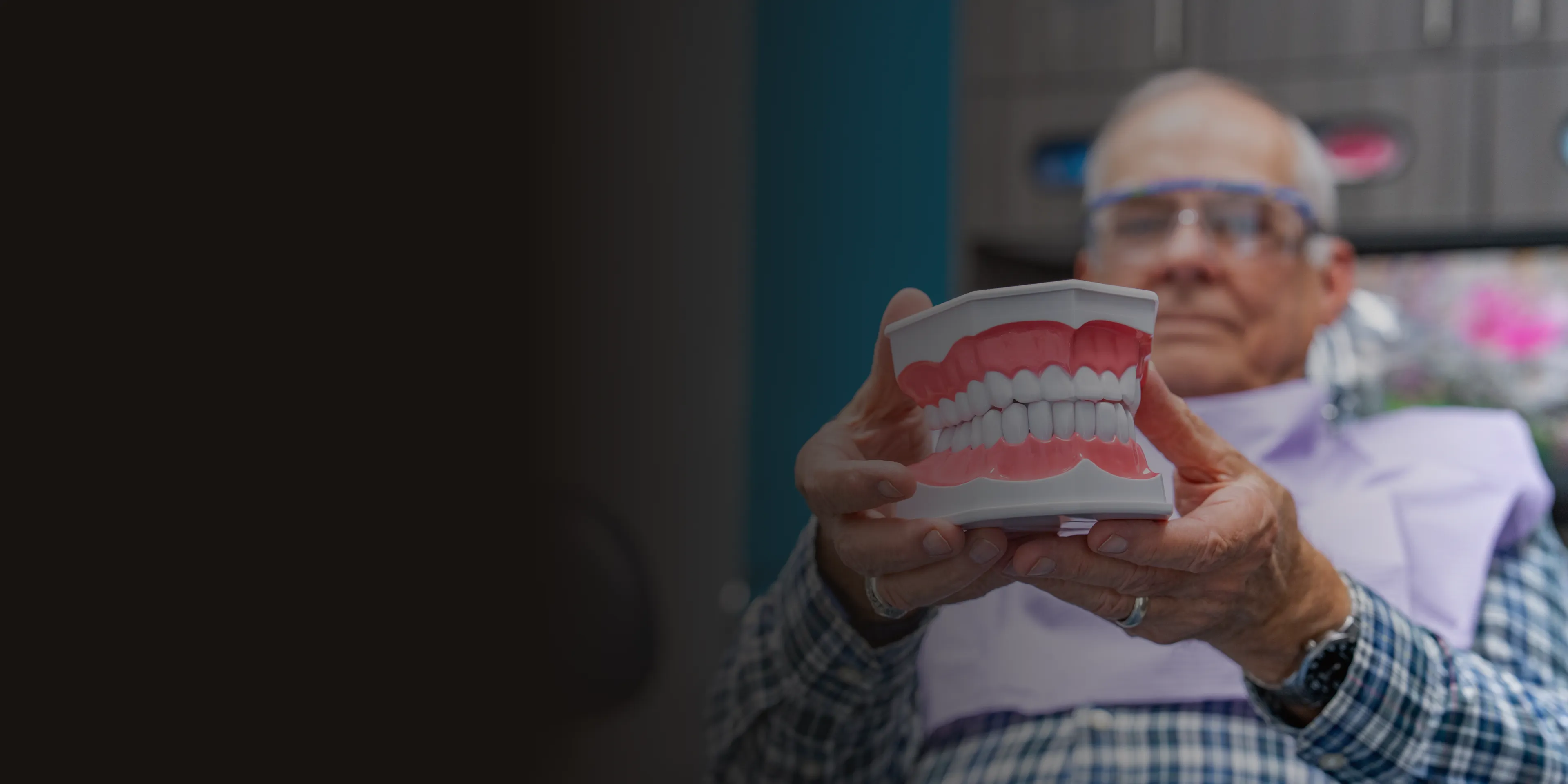 An older man in glasses holds a dental model showing a full set of teeth.
