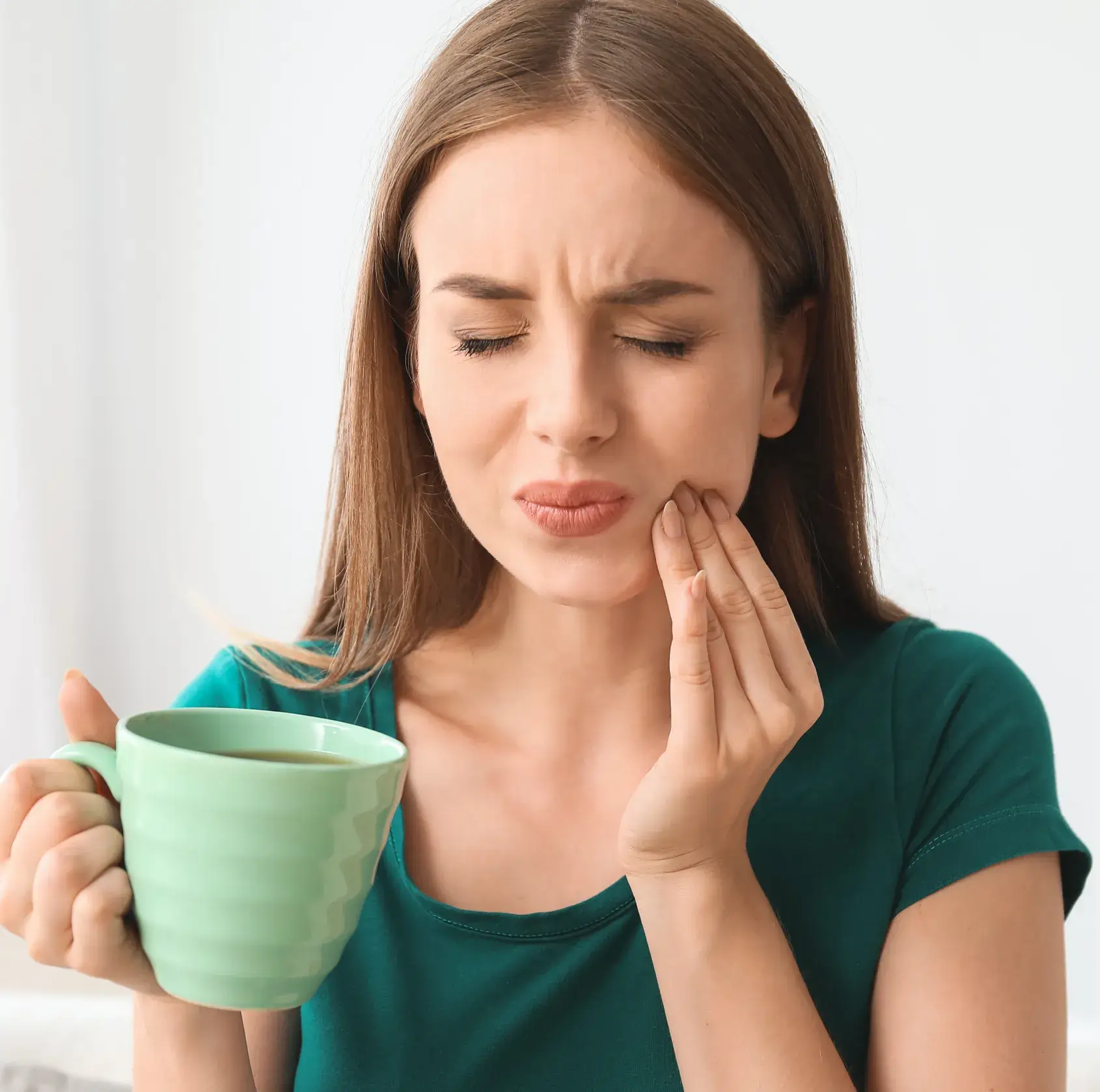 Woman holding a green mug and touching her cheek, appearing to have tooth pain.