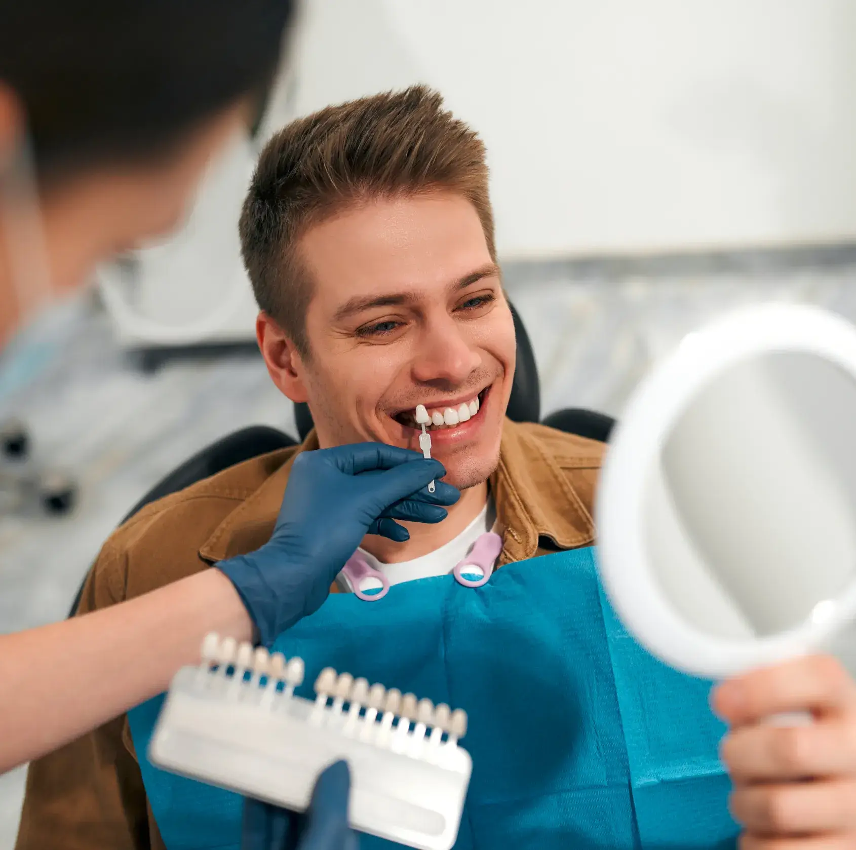 A person is smiling while sitting in a dental chair, holding a mirror during a teeth whitening session.
