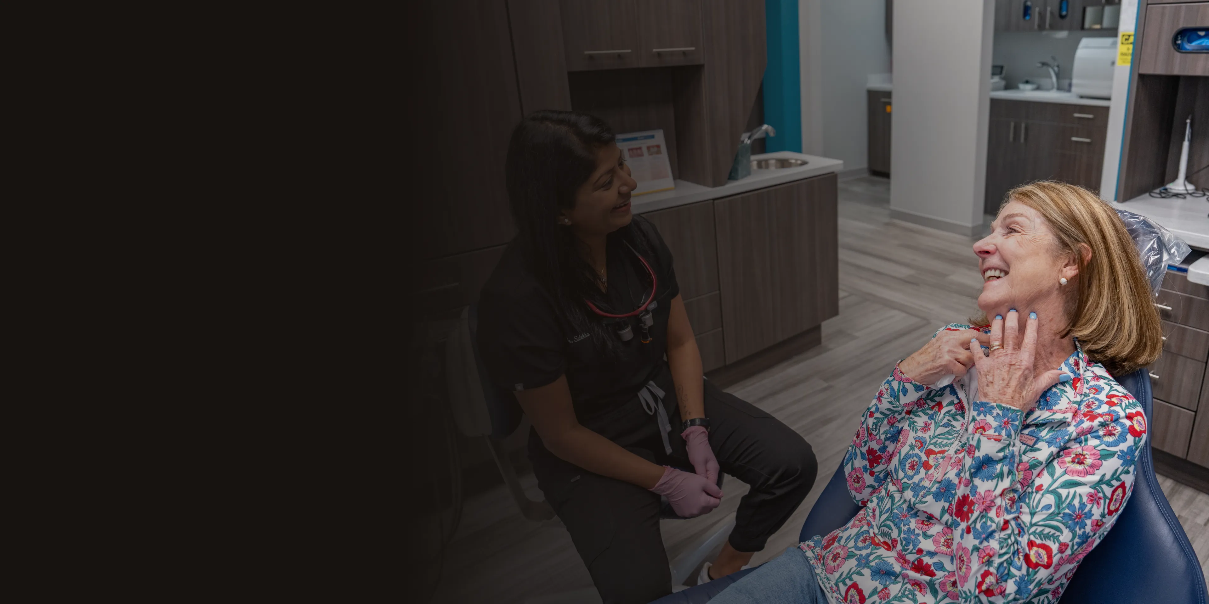 A dentist talks to a smiling patient sitting in a dental chair.
