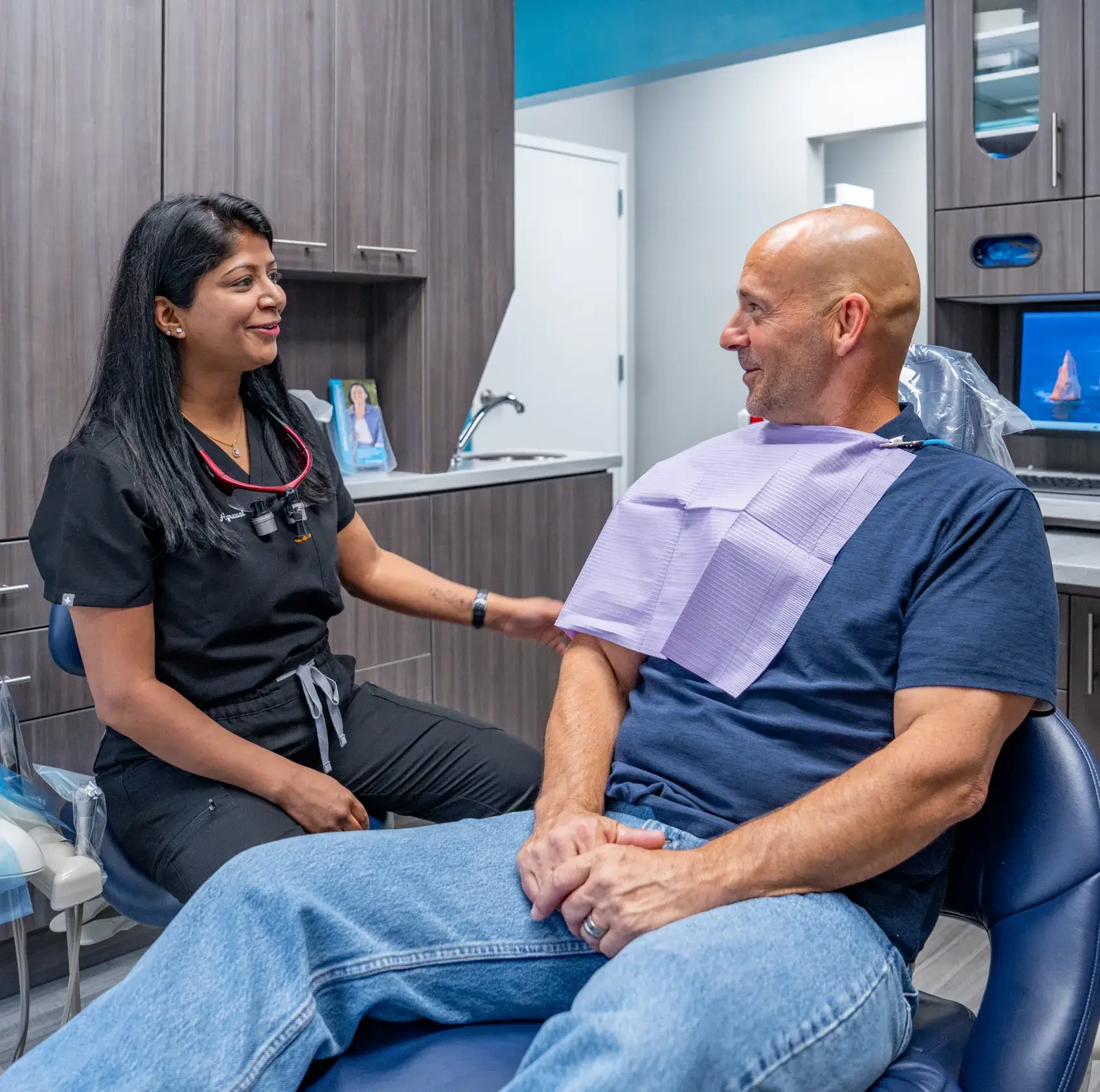 A dentist talks to a patient sitting in a dental chair, creating a friendly interaction.