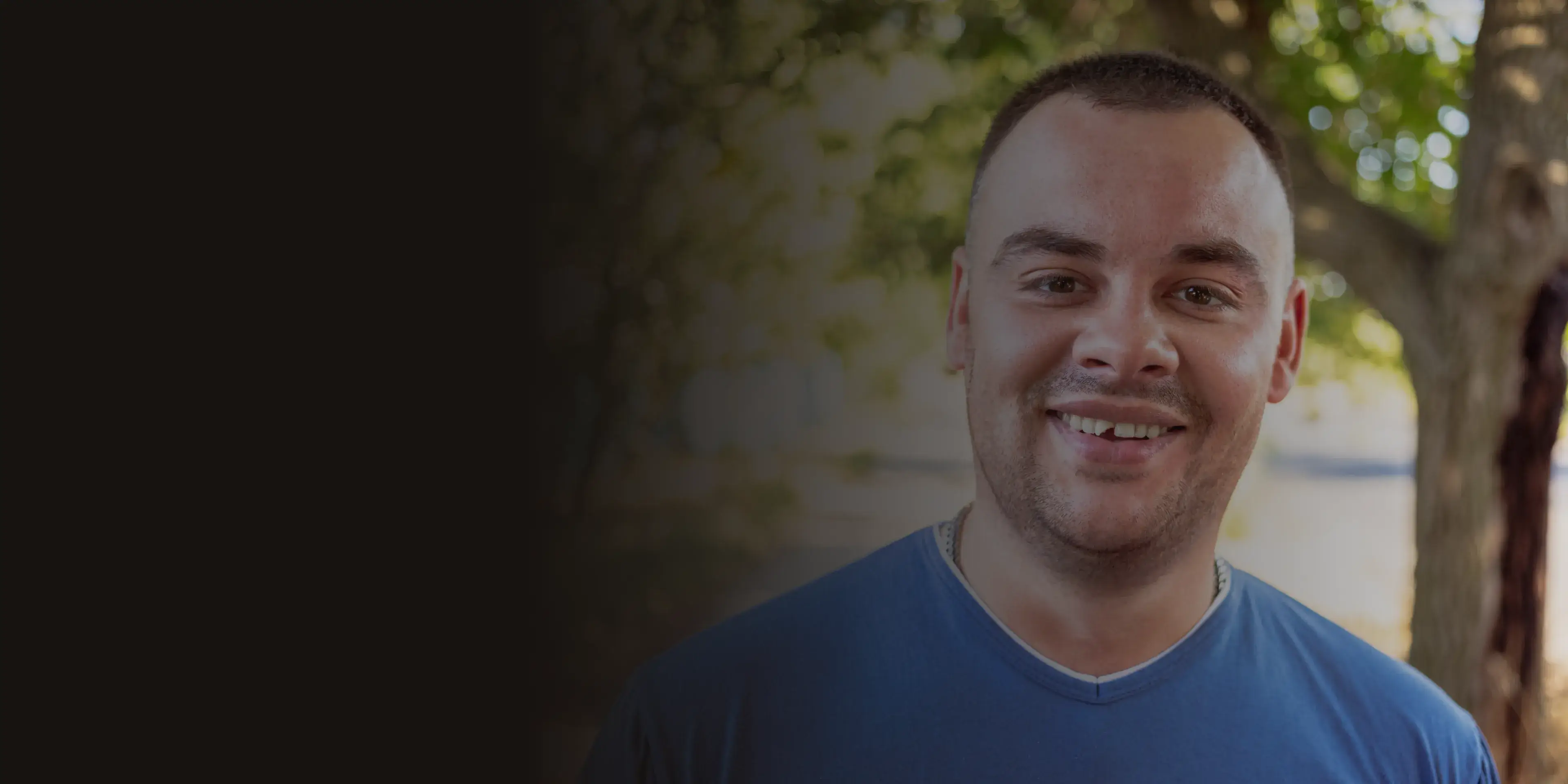 A man with short hair, wearing a blue shirt, smiles outside near trees.