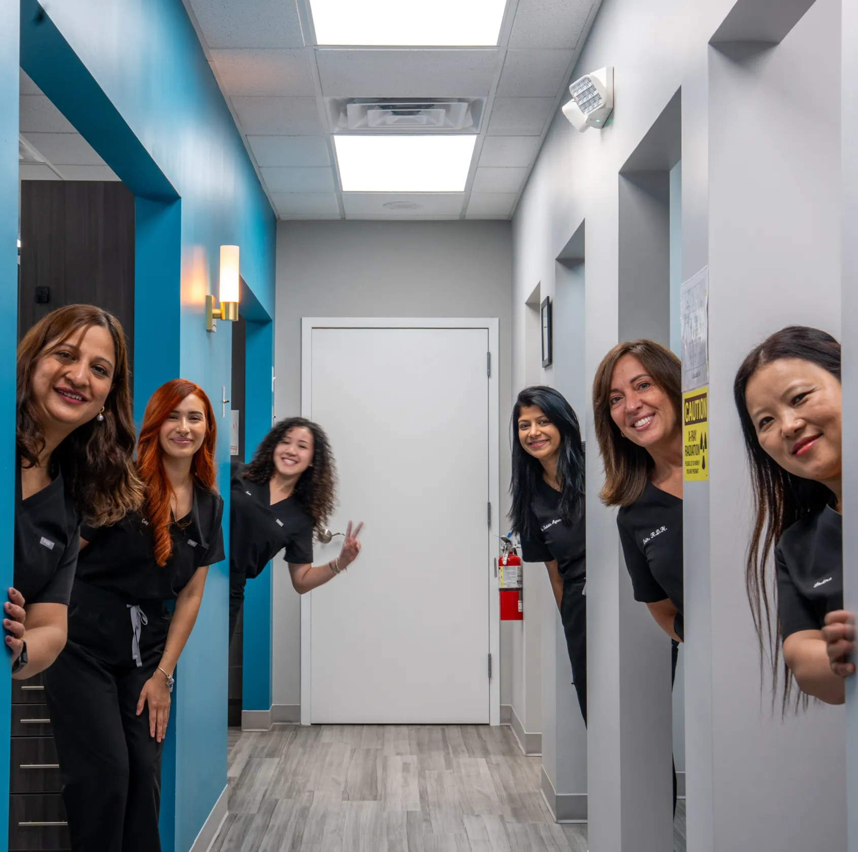 Six smiling women in black uniforms lean out of doorways in a hallway, facing the camera.