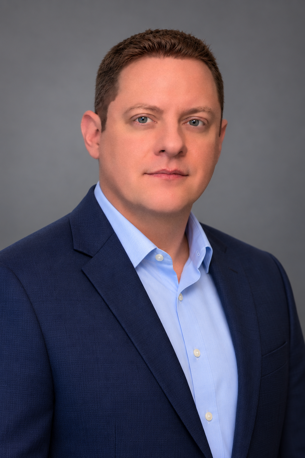 Portrait of Wes Hardcastle wearing a dark blue suit jacket and light blue collared shirt against a plain gray background.