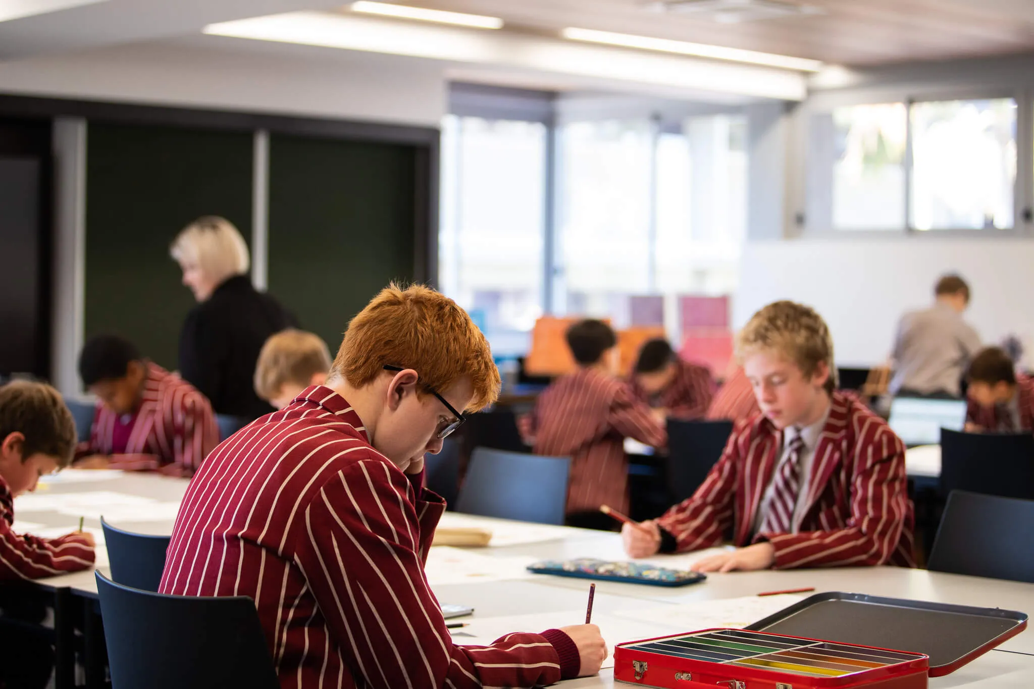 Students in red striped uniforms focused on drawing or writing at desks in a bright classroom.