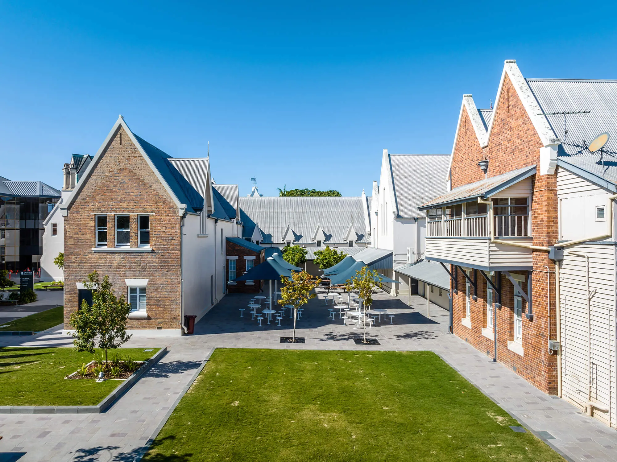 Outdoor courtyard with green lawns and picnic tables shaded by dark blue umbrellas, flanked by brick and white buildings with steep gabled roofs under a clear blue sky.