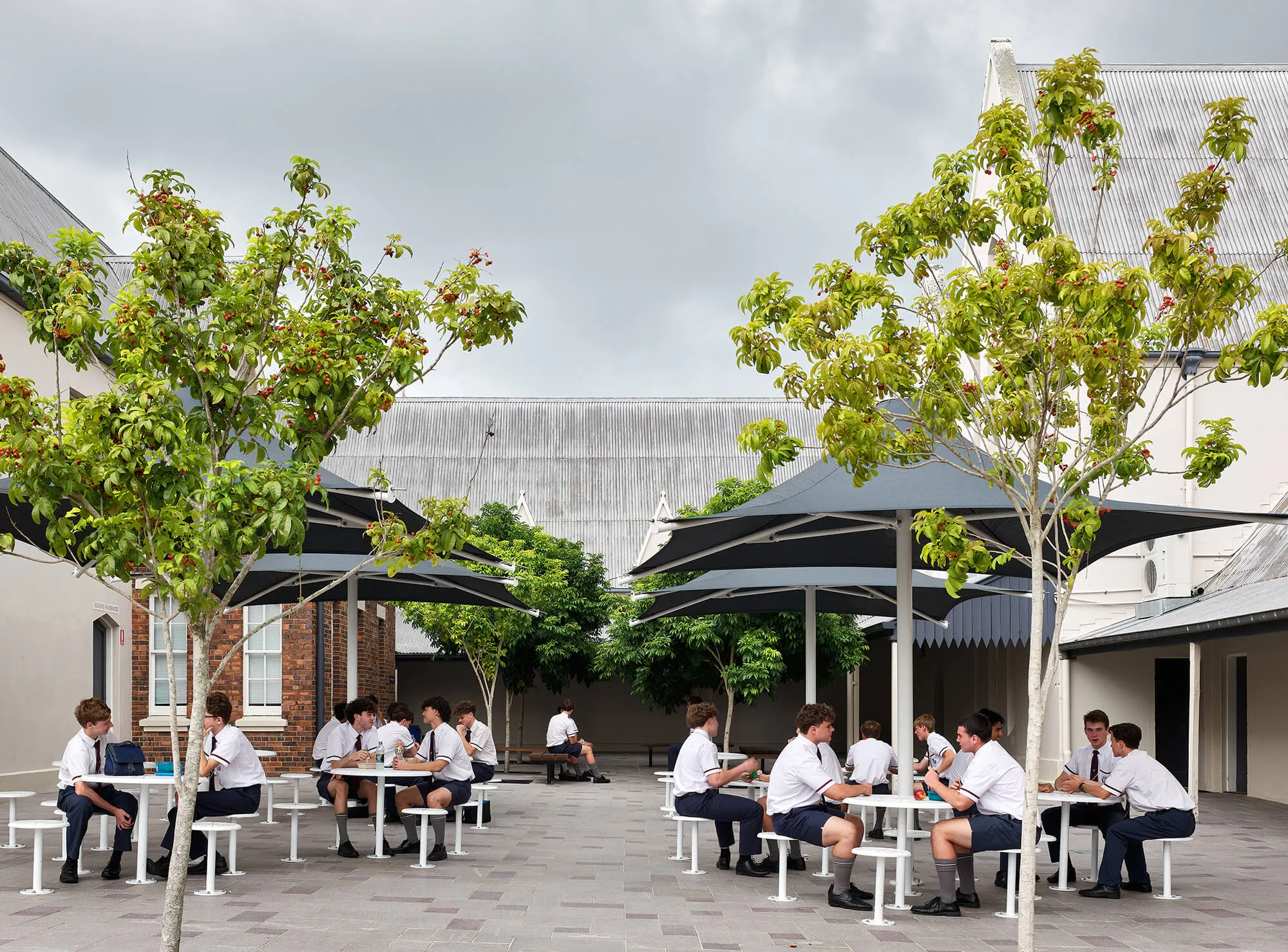 School boys in white shirts and ties sitting and talking at outdoor lunch tables with trees and large umbrellas.