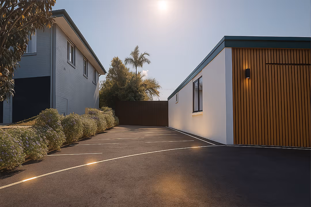 Modern driveway with dark asphalt, parking lines, and landscaped bushes leading to a closed gate between two buildings.