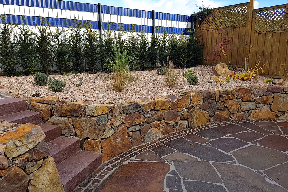 Curved stone retaining wall with mixed rocks, landscaping gravel, plants, and steps next to a paved patio.