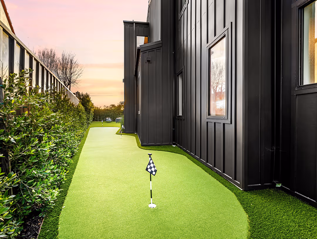 Narrow outdoor putting green with multiple holes next to a modern black building and a hedge fence at sunset.