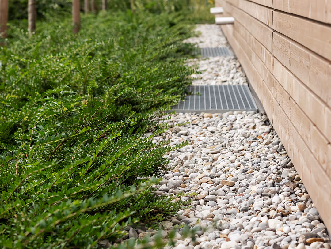 Gravel drainage area bordered by green shrubs alongside a wooden wall with metal grates for water flow.