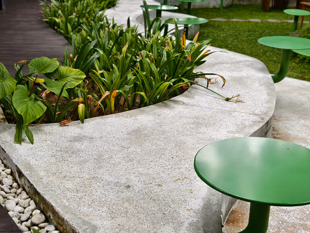 Curved concrete bench surrounding green plants with round green metal tables and green grass in the background.