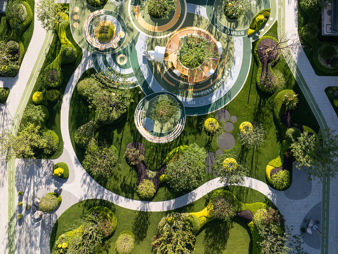 Aerial view of a landscaped urban park with circular pathways, green lawns, various trees and shaded seating areas.