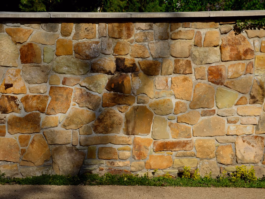 Stone retaining wall with a finished concrete cap, casting shadows on the surface, with greenery above and small plants at the base.