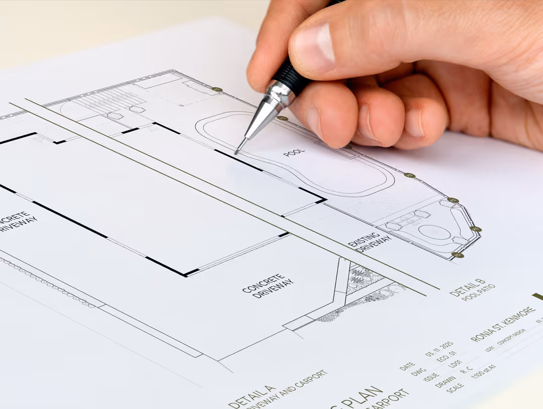 Close-up of a hand holding a pen and drawing on a detailed architectural plan featuring a pool, concrete driveway and carport layout.