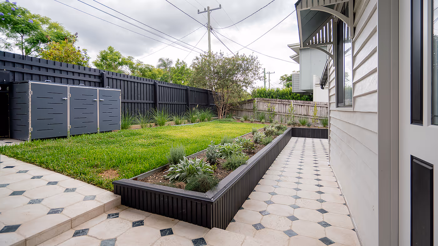 Backyard with raised garden bed, green lawn, and tiled patio beside a white house under a cloudy sky.