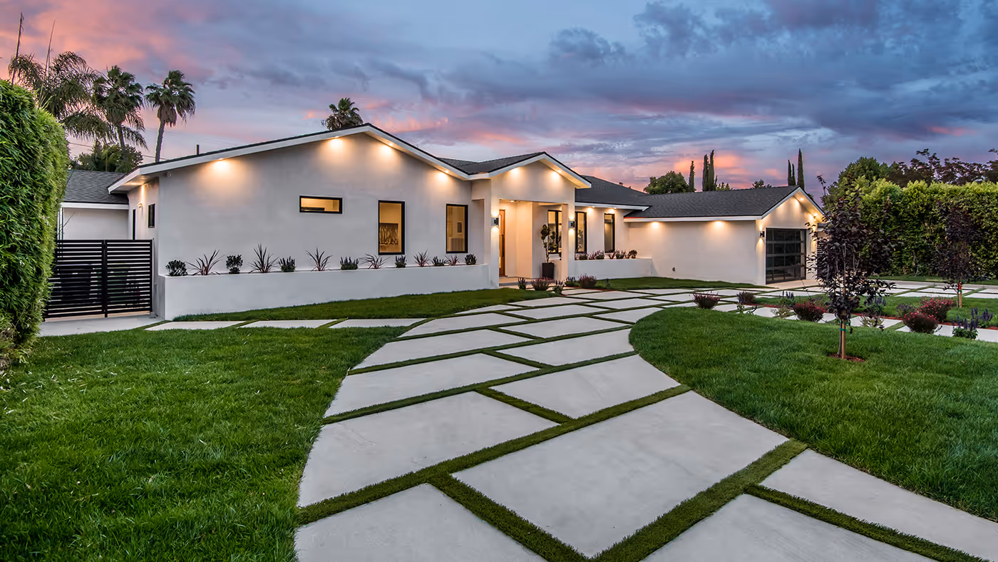 Modern single-story house with illuminated exterior lights, geometric concrete driveway with grass inlays, green lawn, and evening sky.