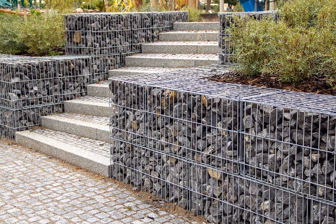 Outdoor stone steps and retaining walls constructed with metal cages filled with dark rocks, surrounded by paved stone ground and greenery.