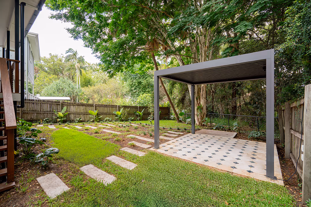 Backyard with green grass, stepping stone pathway, and a modern grey pergola on a tiled patio, surrounded by trees and fence.