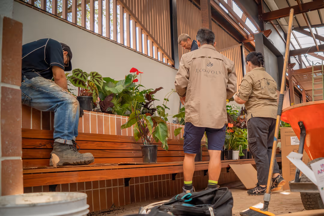 Three Ecovolve team members are arranging potted plants on a wooden bench, with gardening tools and equipment nearby.