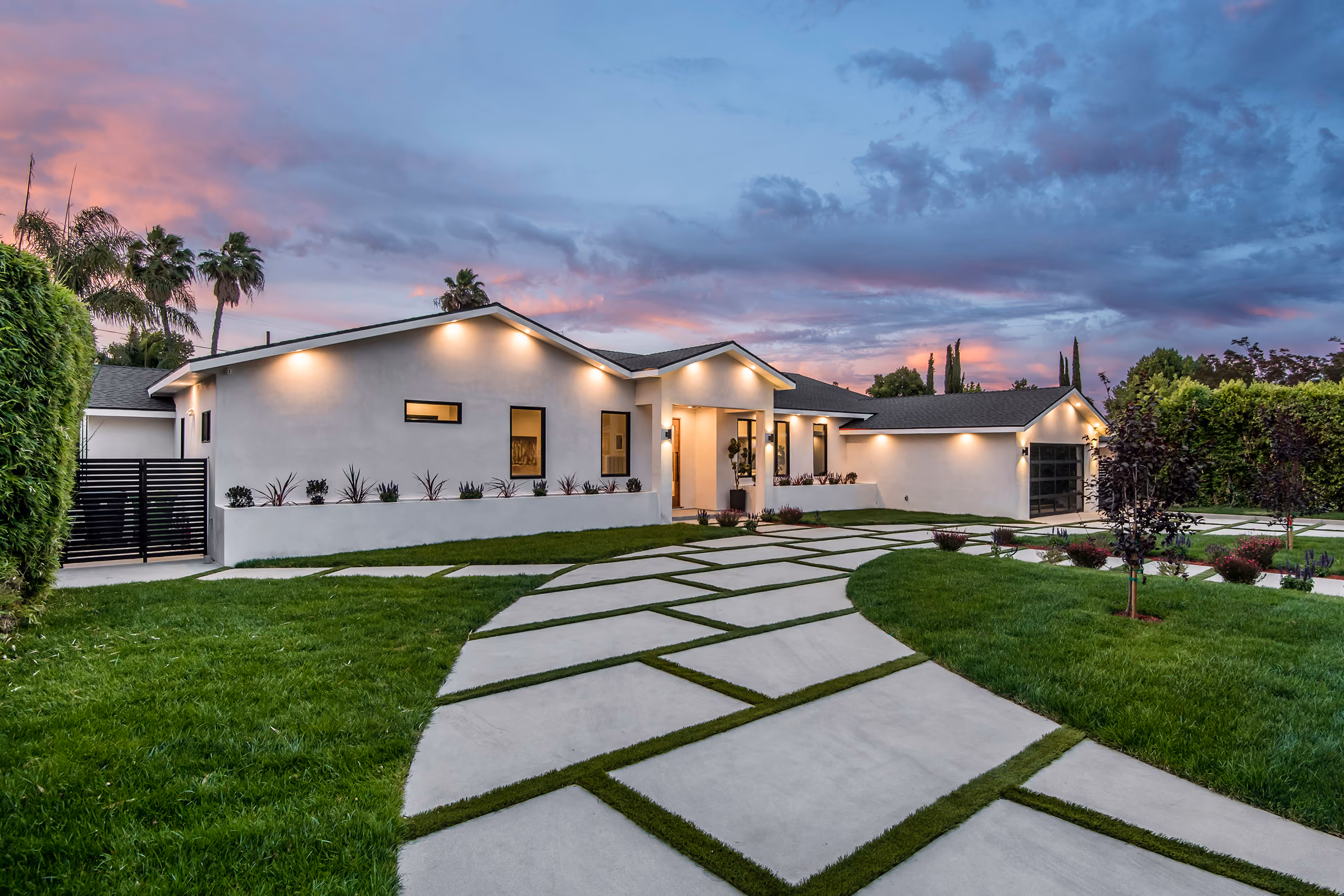 Modern single-story house with illuminated exterior lights, geometric concrete driveway with grass inlays, green lawn, and evening sky.