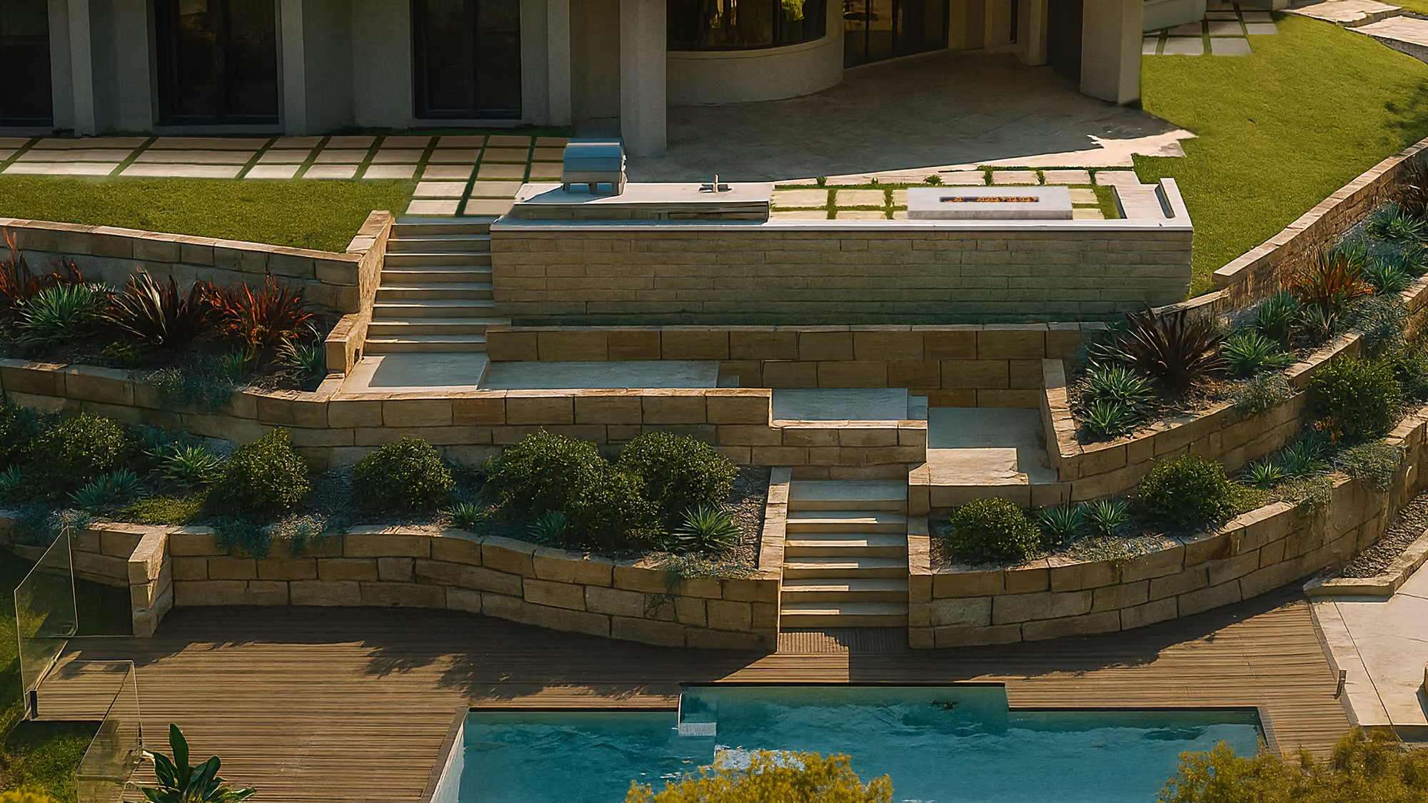 Modern terraced backyard with stone retaining walls, greenery, stairs, a wooden deck and a swimming pool.