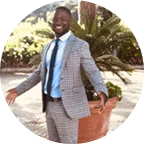 Lanre Olaitan, founder of PriveDoc, smiling and posing in a grey checkered suit outdoors in front of a potted plant.