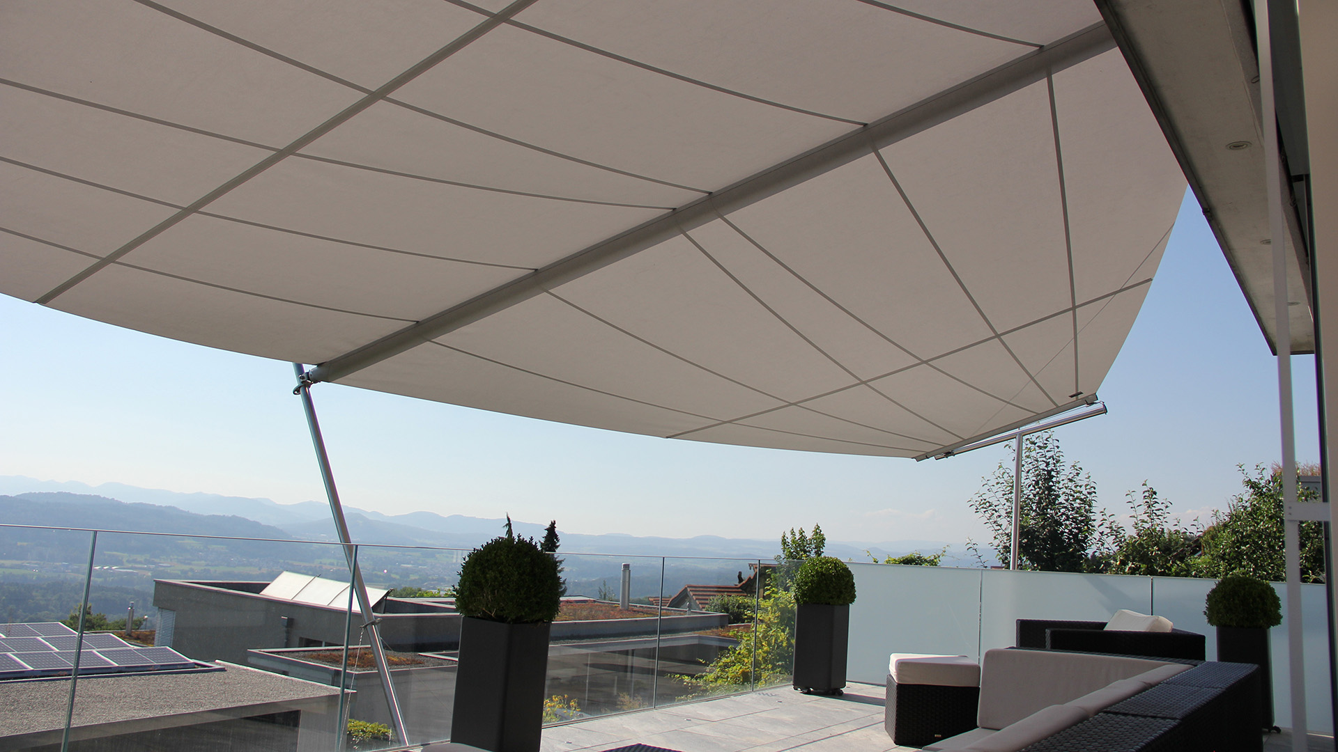 Eine stützenfreie Terrasse mit einem trapezförmig zugeschnittenen SunSquare Segel mit 33 m² in Sattler 321-030 Hellgrau als ZickZack Segel gefertigt. Die Terrasse mit Weitblick erfährt Schutz vor Sonne und Regen.