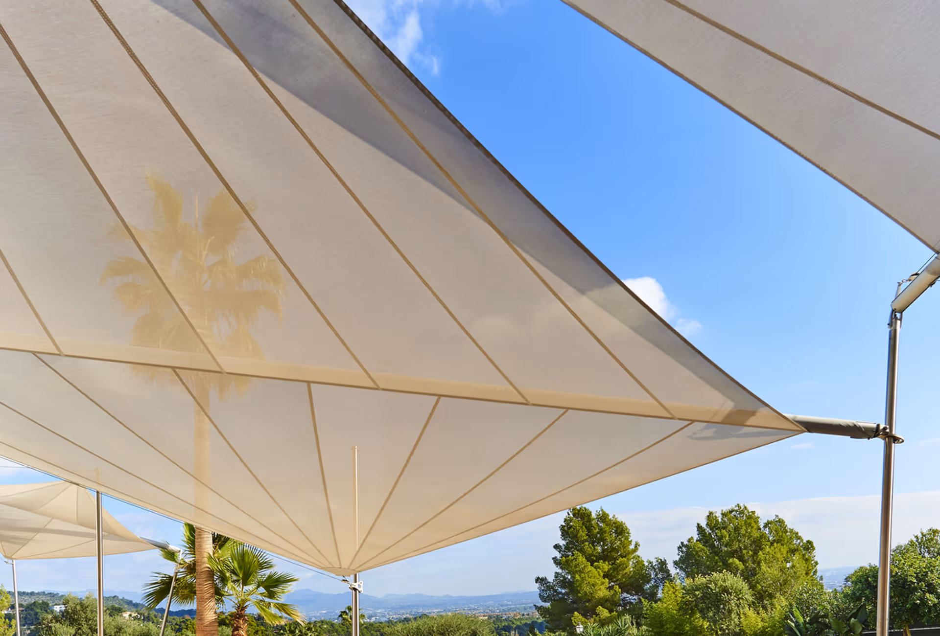 Eine Nahaufnahme zeigt ein SunSquare Sonnensegel, gefertigt aus Serge Ferrari Soltis 92, als reiner Sonnenschutz, daher auch die sanfte Transparenz des Segels - man sieht die Palme im Hintergrund durch das Segel scheinen.