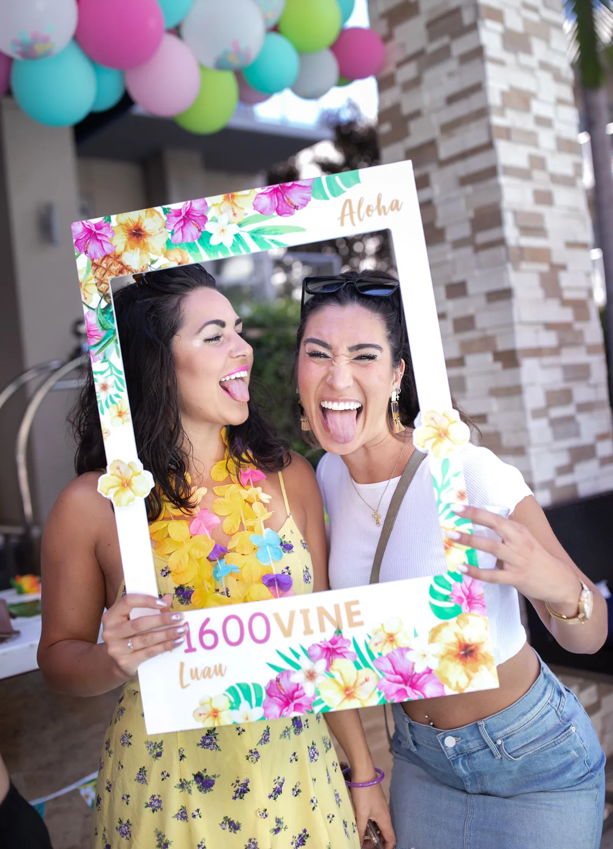 Two people posing with a cardboard frame labelled "Aloha 1600 VINE Luau".