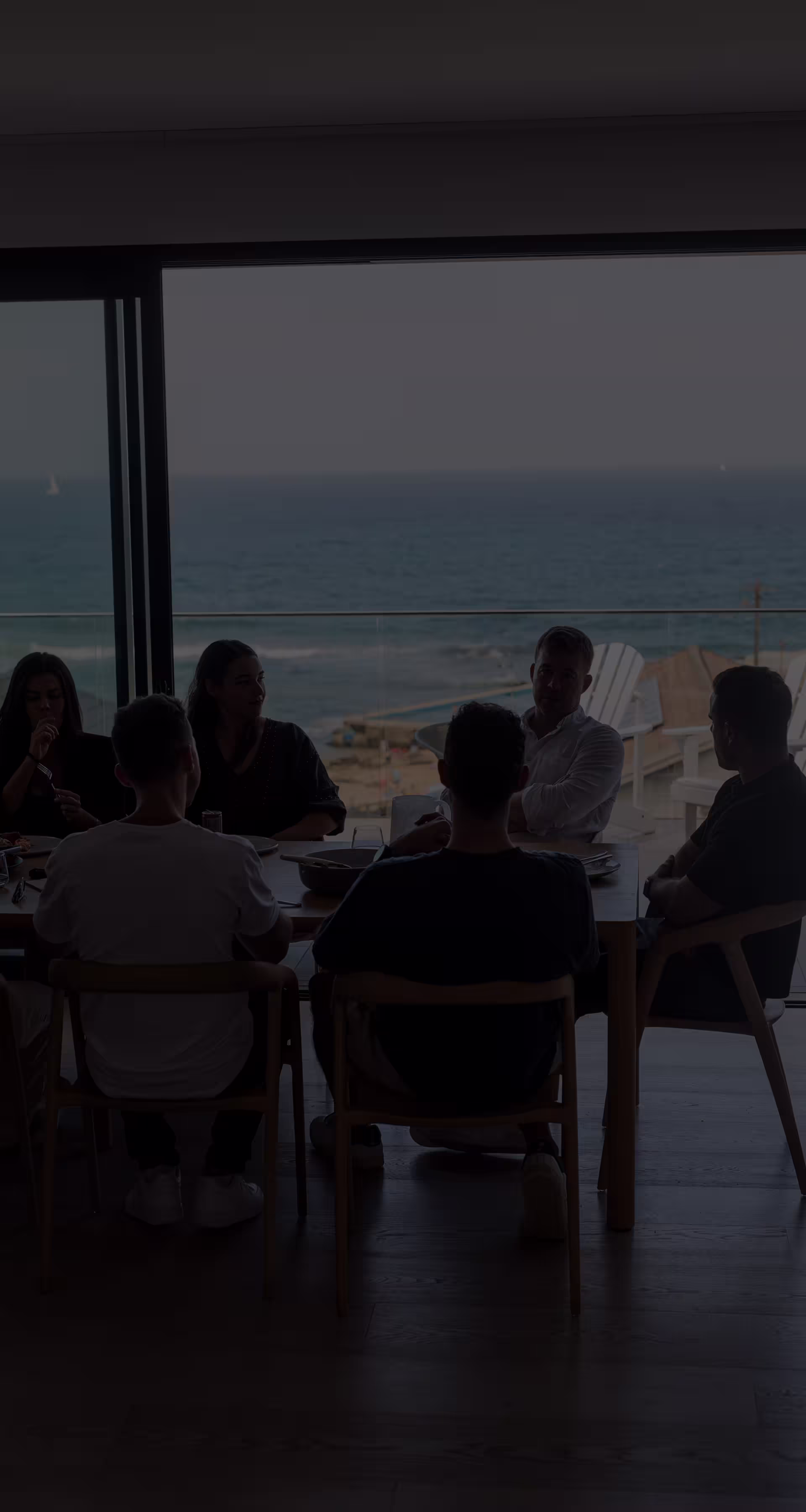 A group of people sitting around a table indoors, with windows showing an ocean view in the background.